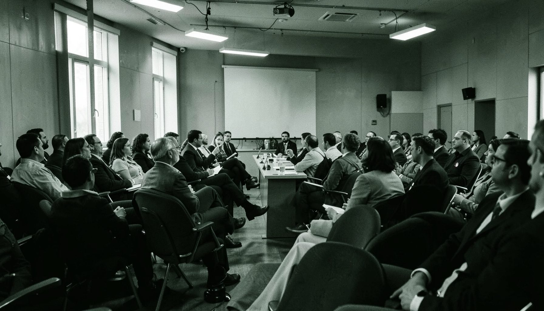 Fotografia in duotone bianco e nero di una sala conferenze con persone sedute in ascolto, atmosfera raccolta