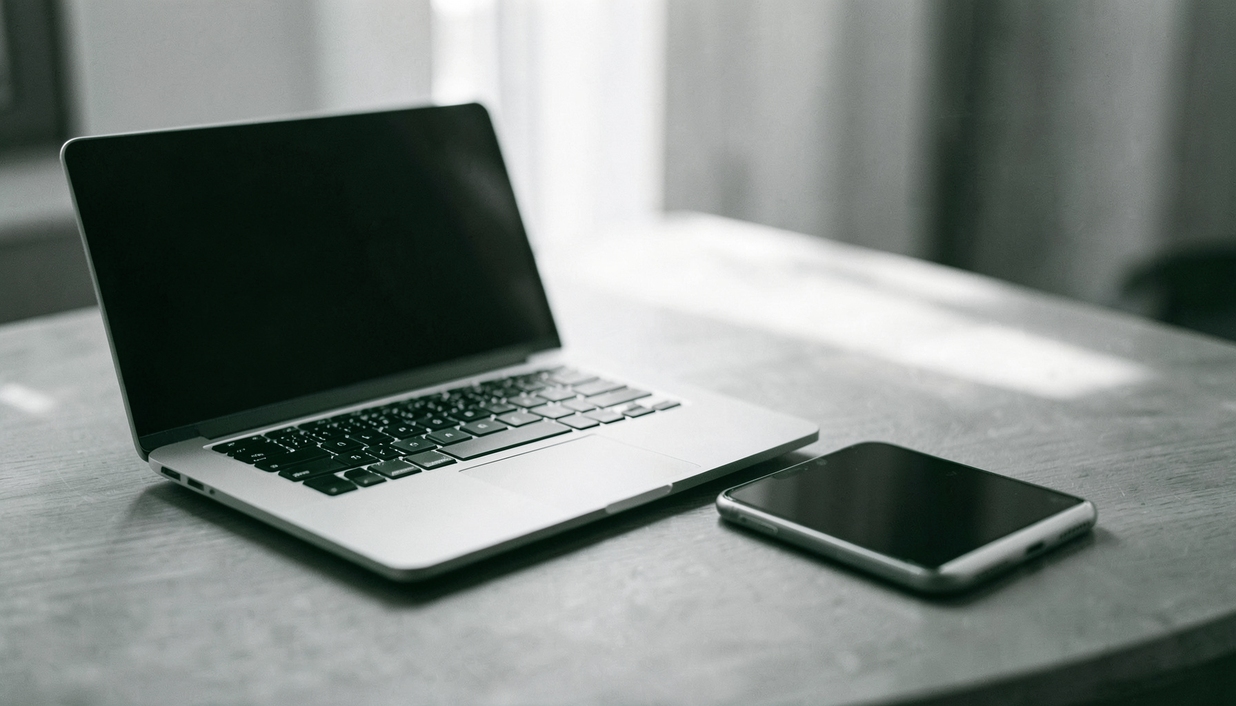 Fotografia in duotone bianco e nero di un laptop e uno smartphone appoggiati su una scrivania, luce naturale soffusa
